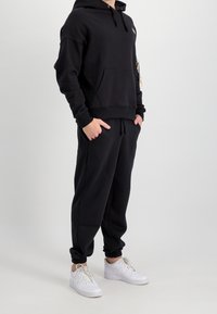 Felpa nera con cappuccio e tasca frontale, abbinata a pantaloni joggers neri. Sneakers bianche completano l'outfit. Tessuto morbido e liscio.