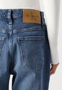Calvin Klein zili džinsi ar aizmugures kabatām, bežīgas ādas logotipa uzšuvums un redzami šuvju elementi. Raksturīgs taisnā kāju dizains.