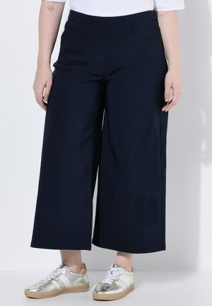 Frau trägt schwarze, weit geschnittene, kurze Hosen und metallisch-silberne Sneaker, steht vor einem schlichten weißen Hintergrund.