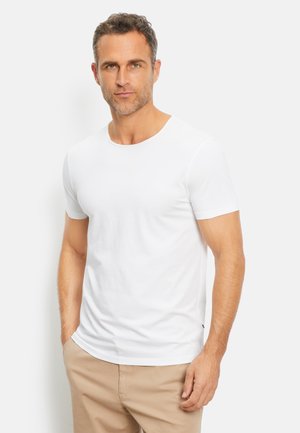 Uomo con capelli corti e ricci che indossa una maglietta bianca a maniche corte semplice e pantaloni beige, in piedi su uno sfondo bianco.