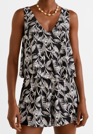 Femme portant une combinaison noire sans manches à motif de feuilles blanches et col en V, accessoirisée avec un collier de coquillages.