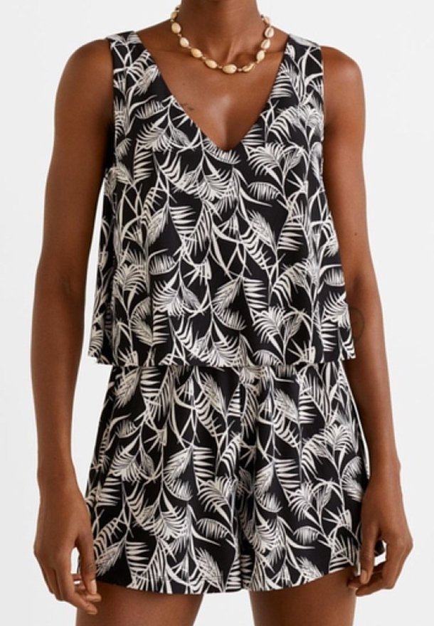 Femme portant une combinaison noire sans manches à motif de feuilles blanches et col en V, accessoirisée avec un collier de coquillages.