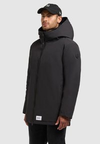Parka nera isolata con colletto alto e cappuccio staccabile. Presenta una cerniera frontale e un patch con logo sulla manica sinistra. Texture liscia.
