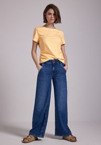 Camiseta amarilla con estampado y mangas cortas, combinada con jeans anchos de color azul. El atuendo incluye sandalias marrones claras con detalles metálicos.