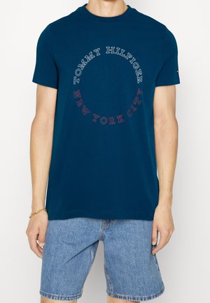 Osoba nosząca granatowy T-shirt z okrągłym napisem „Tommy Hilfiger New York City” oraz jasnoniebieskie szorty jeansowe.