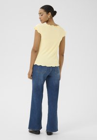 Haut jaune à côtes avec des bords festonnés, associé à un jean bleu foncé évasé taille haute. Une silhouette lisse et ajustée avec une texture légère.
