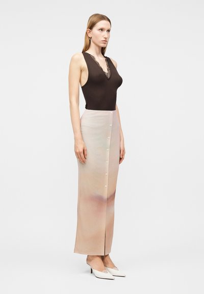 Γυναίκα με ίσια ανοιχτό καστανά μαλλιά που φοράει μαύρο αμάνικο τοπ με δαντέλα, μπεζ μανικετόφουστα με κουμπιά και λευκά παπούτσια με τακούνι.