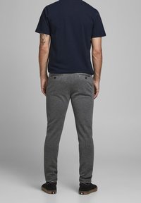 Šedé tepláky s tapered střihem, vyrobené z měkkého materiálu. Mají dvě zadní kapsy a černou obuv s hnědou podrážkou. Jsou vidět modré tričko.