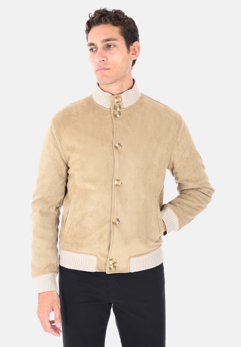 Uomo in giacca scamosciata beige con colletto e polsini in maglia beige, pantaloni neri, una mano in tasca, che guarda di lato su sfondo bianco.