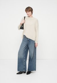 Γυναίκα που φοράει μια κρεμ ζακετοφόρα μπλούζα, φαρδιά μπλε τζιν παντελόνια, μαύρες μπότες μέχρι τον αστράγαλο και κρατάει μια μικρή μαύρη τσάντα στον ώμο.