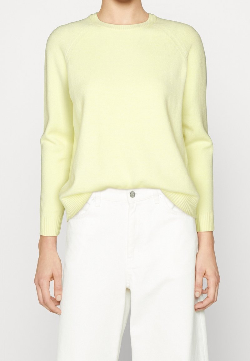 Personne portant un pull jaune clair à manches longues rentré dans un pantalon blanc taille haute sur un fond uni.