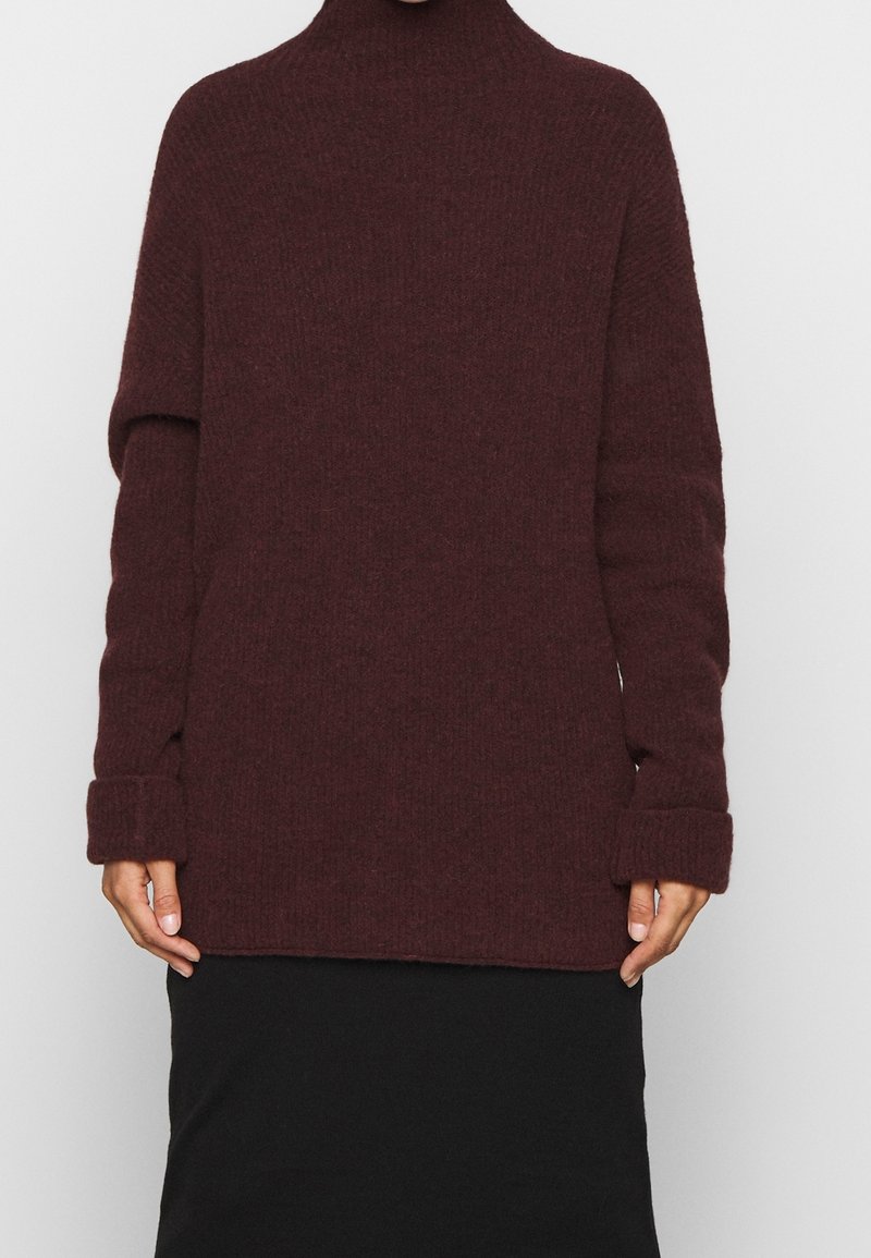 Pull en tricot bordeaux avec un col montant, une coupe oversize et des poignets pliés, associé à une jupe noire. Texture douce partout.