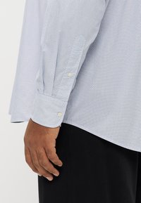 Vaaleansininen kuvioitu nappipaita, jossa on pitkät hihat ja napilliset hihansuut, näyttää teksturoitua kangasta ja rentoa istuvuutta. Mustat housut näkyvissä.