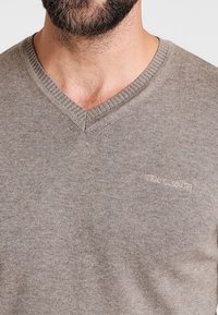 V-ringad tröja i ljusbrun stickad tyg med ribbad krage. Har en broderad logotyp på vänster bröst. Mjuk textur, avslappnad design.