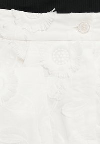 Λευκή φούστα με κεντημένα λουλούδια και υφασμάτινη υφή, διαθέτοντας λείο ζωνάκι και κλείσιμο με κουμπί, αναδεικνύοντας περίπλοκες λεπτομέρειες ραφής.