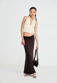 Beige halter crop top met een geplooide buste, gecombineerd met een aansluitende bruine maxirok. Model houdt een zwarte handtas vast en draagt lichte sandalen.