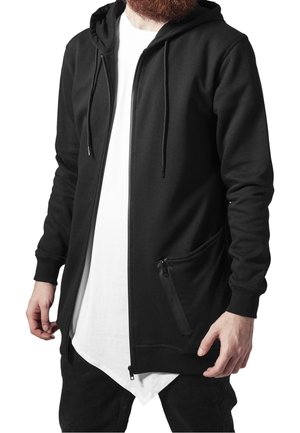 Mężczyzna ubrany w czarną bluzę z kapturem na zamek błyskawiczny z przednią siatkową kieszenią, na białej, dłuższej koszulce oraz czarnych spodniach.