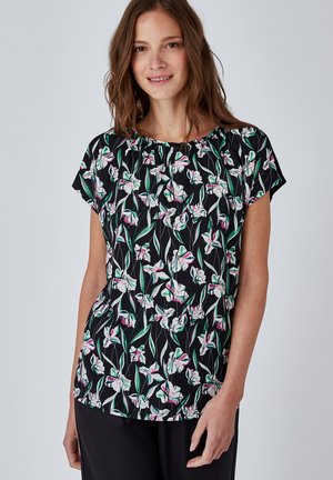 Donna che indossa una camicetta nera a maniche corte con motivo floreale rosa e bianco e pantaloni neri, sorridente con capelli castani sciolti.
