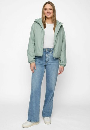 Jeune femme debout, portant une veste à capuche vert clair, un t-shirt blanc, un jean taille haute bleu et des baskets blanches sur fond blanc.