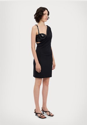 Zwarte jurk met een one-shoulder ontwerp, gedrapeerde stof en een glanzende zwarte beha-achtige top. Zwarte gekruiste hakken maken de look compleet.