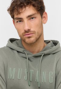 Hoodie vert olive avec cordons ajustables, présentant un logo "MUSTANG" en relief bien en vue dans une couleur assortie. Tissu doux et texturé.