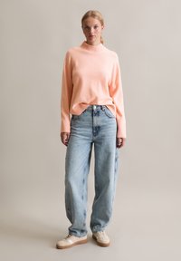 Maglione rosa chiaro con colletto alto abbinato a jeans azzurri a vestibilità rilassata e sneakers crema. Texture liscia con dettagli a maglia sottili.