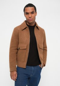 Giacca di lana marrone con un colletto, due tasche frontali e cerniera argentata. Indossata sopra un maglione nero, abbinata a jeans blu.