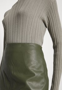 Grå ribstrikket sweater under et tætsiddende mørkegrønt lædernederdel. Teksturerne kontrasterer, hvilket fremhæver læderets glathed.