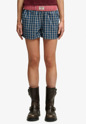 Osoba nosząca niebiesko-białe kraciaste szorty z czerwoną gumką w pasie, bordową koszulkę oraz ciemnobrązowe skórzane botki do połowy łydki z klamrami.