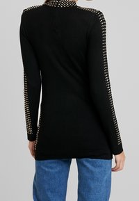 Top negro de manga larga con cuello alto, que presenta detalles de tachuelas doradas a lo largo de las mangas y un diseño ajustado.