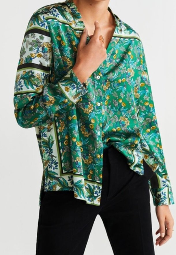 Personne portant une chemise boutonnée à manches longues à motif floral vert avec un pantalon noir, tenant le col de la chemise d'une main.