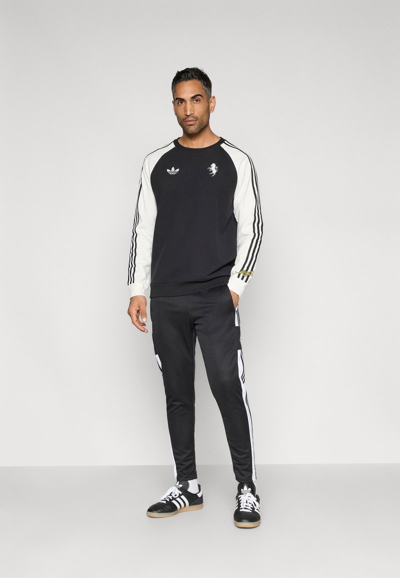 Schwarz-weißes Sport-Sweatshirt mit langen Ärmeln, das ein Logo auf der Brust und dreistreifige Details zeigt. Kombiniert mit schwarzen Jogginghosen.