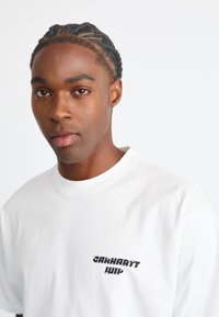 Witte katoenen t-shirt met een zwarte logoprint op de linkerborst, korte mouwen en een ronde hals. Soepele textuur.