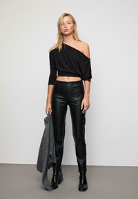 Crna skraćena bluzica s otkrivenim ramenima, prilagođene crne kožne hlače i crne gležnjače. Model drži sivi džemper. Glatke teksture i elegantan dizajn.