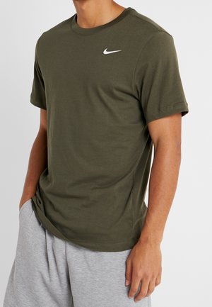 Mand iført en olivengrøn Nike kortærmet t-shirt og lysegrå shorts, stående med den ene hånd i lommen mod en ensfarvet baggrund.