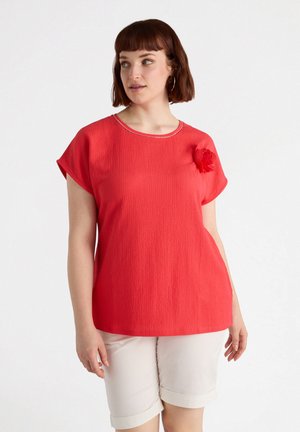 Top rosso con texture, scollatura a girocollo, maniche a pipistrello e un accento floreale sulla spalla. Abbinato a pantaloni corti bianchi con risvolto.