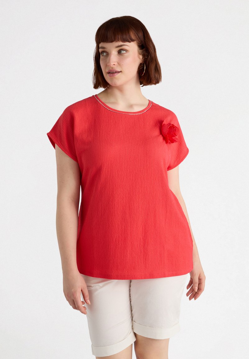 Top rojo texturizado con escote de barco, mangas cortas y un acento floral en el hombro. Combinado con pantalones cortos blancos con puños.