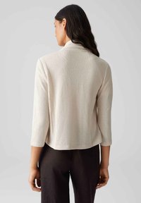 Beige strukturierte Jacke mit drei Viertelärmel, offenem Front und Kragen. Trägt über dunklen Hosen und zeigt eine lässige Silhouette.