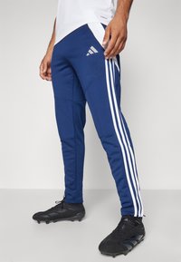 Blaue sportliche Hosen mit weißen Seitenstreifen und einem Adidas-Logo auf dem Oberschenkel, aus einem glatten, flexiblen Stoff gefertigt. Schwarze Stollenschuhe.