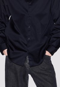 Camicia blu navy con finitura liscia, dotata di un logo sul petto, maniche lunghe e orlo curvo, abbinata a jeans scuri con texture.