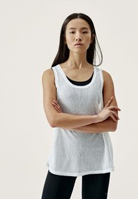 Vit ärmlös topp med ett texturerat mönster, rund halsringning och avslappnad passform, lager på lager över en svart tanktop. Matchat med svarta leggings.