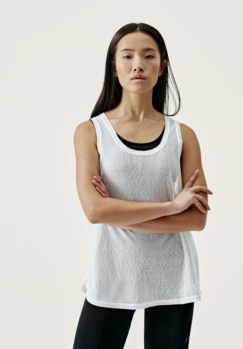 Vit ärmlös topp med ett texturerat mönster, rund halsringning och avslappnad passform, lager på lager över en svart tanktop. Matchat med svarta leggings.
