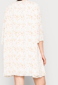 Femme portant une robe plissée blanche avec un motif floral orange clair, vue de dos avec un bras détendu à ses côtés.