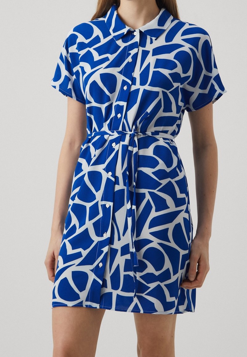 Femme portant une robe à manches courtes, boutonnée, avec des motifs géométriques abstraits bleus et blancs et une ceinture nouée à la taille.