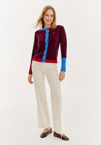 Bordowy kardigan z niebieskim kołnierzykiem i guzikami, czerwonym pasem, długimi rękawami z niebieskimi mankietami, zestawiony z beżowymi spodniami i wzorzystymi balerinami.