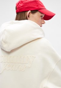 Cremefarbene Kapuzenjacke mit gesticktem "Tommy Jeans"-Logo, kombiniert mit einer roten Mütze. Weicher Stoff und entspannter Schnitt. Die Kapuzenjacke verfügt über eine geräumige Kapuze.