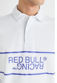Balta polo krekla ar divu pogu aizdari, mīkstu tekstūru un izteiksmīgu zilo "RED BULL RACING" logo priekšā.