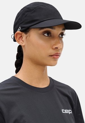 Woman with braided hair wearing a black cap and dark gray athletic shirt with "cep" logo, facing right in front of a neutral background.