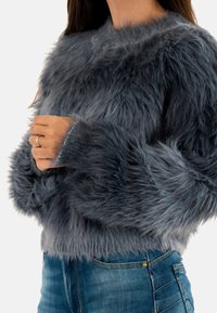 Suéter de piel sintética en gris oscuro con mangas largas y esponjosas, escote redondo y una textura suave, combinado con vaqueros de denim azul.