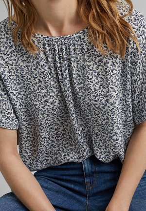 Femme portant un chemisier blanc avec de petits motifs de feuilles bleues et un jean bleu, assise les mains posées sur les cuisses.